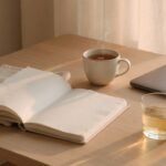 Morning focus desk with open notebook, ceramic cup, glass and closed laptop in soft light